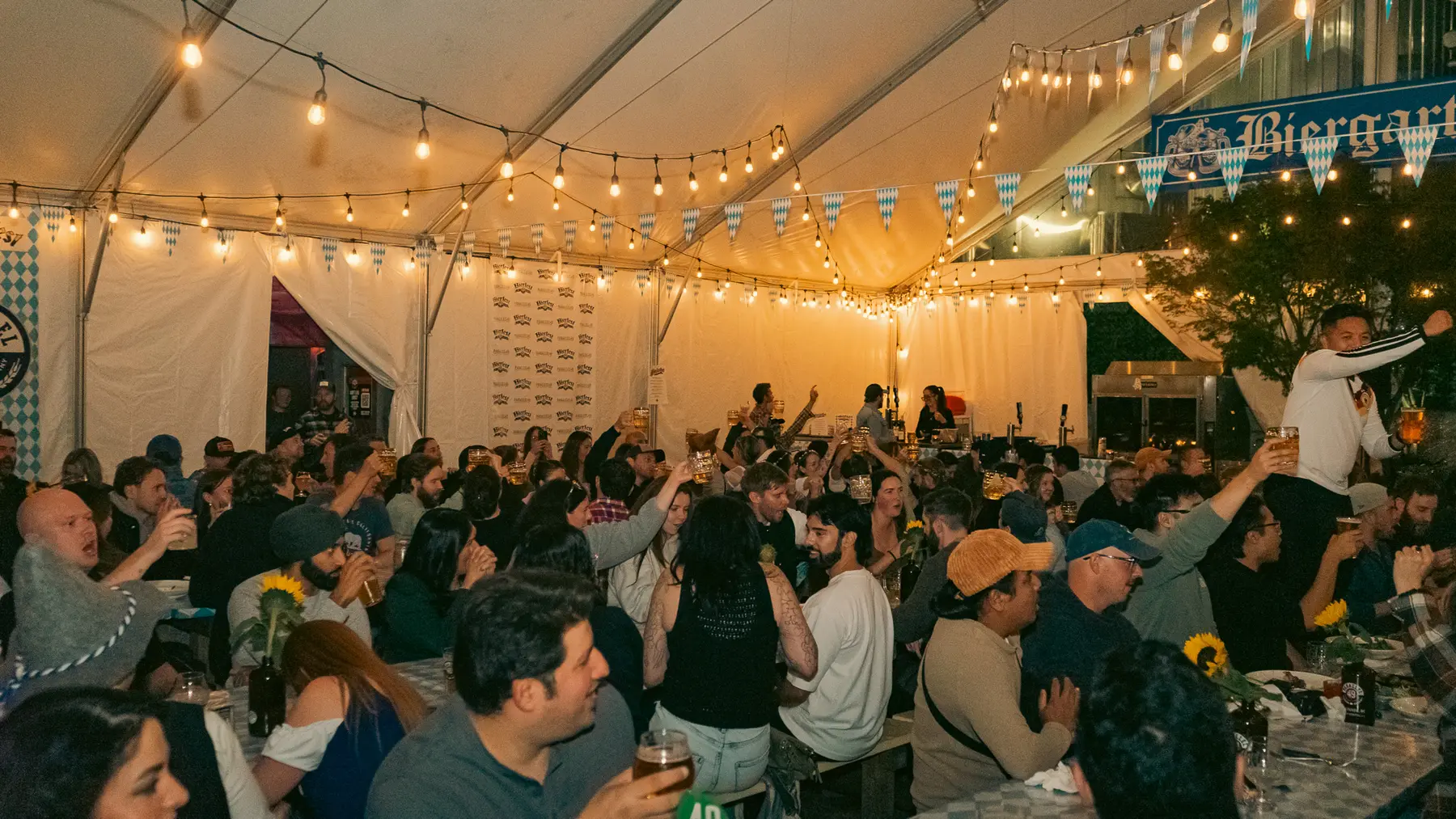 A packed Oktoberfest biergarten tent at Parallel 49 under string lights and Bavarian bunting, guests raising steins