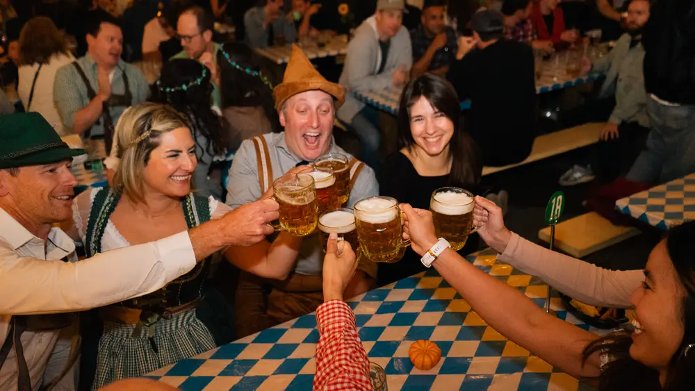 Guests in Bavarian hats and dirndls clinking beer steins over a blue-and-white checked table